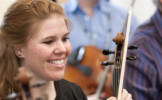 A red haired woman smiling and holing a violin and bow
